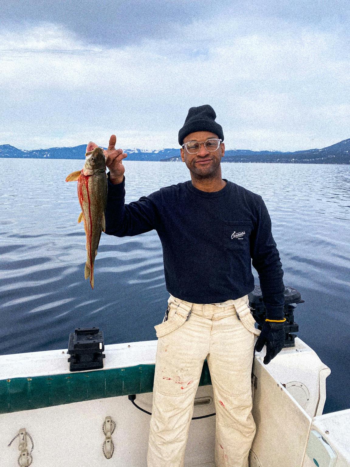 A person with glasses holds up a fish with one hand while standing on a boat.